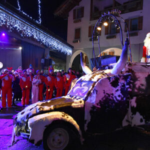 Christmas Chalets in the Alps Santa Claus waves from a snow-dusted, horned parade car as a group in red ski suits gathers beside a building draped in icicle lights.