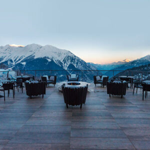 European ski escapes for a luxury weekend getaway An empty mountain terrace with wicker armchairs and a central fire pit overlooks snow-capped peaks at dusk.