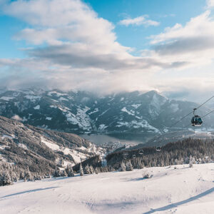 Discover the diverse resort of Zell am See-Kaprun Snow-covered mountain slope with cable cars suspended on wires above a pine forest and lake, beneath a partly cloudy sky.