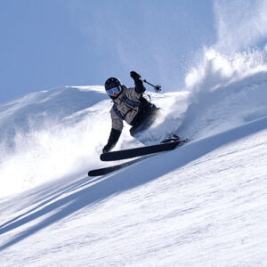 A skier carves through deep powder on a sunlit slope, sending up a dramatic spray of snow against a clear blue sky.