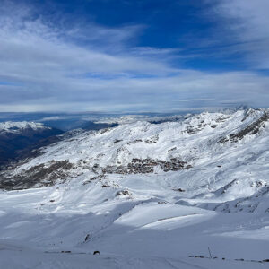 Snow-covered alpine mountains stretch into the distance with a small village nestled in the valley and a ski lift on the left.