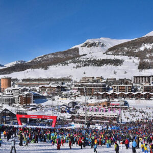 Vialattea to host additional Giant Slalom race A large crowd of skiers gathers on snow below a mountainside village, with snow-capped peaks under a clear blue sky.
