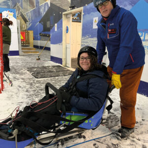 A smiling skier in a helmet sits strapped into a blue sit-ski on snow, helped by a person in a blue jacket.
