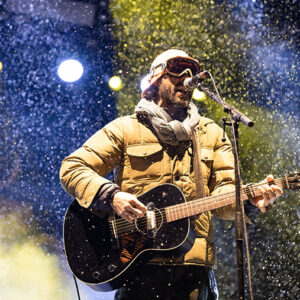 Rendezvous Music Festival Returns to Jackson Hole this April A bearded performer in a cap, scarf and tan puffer jacket plays a black acoustic guitar and sings into a microphone as snow falls through bright stage lights.