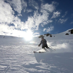 Skier to attempt new World record – 100 different ski lifts in one day Skier carving through powder on a sunlit slope beneath a bright, cloud-streaked sky.