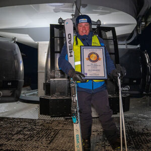 Skier sets new world record with 101 ski lift rides in a day Smiling man in a hi-vis vest and helmet holds HEAD skis and a framed certificate between numbered gondola cabins at night.