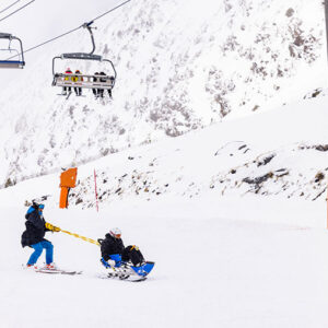 A skier tows a sit-ski down a snowy slope beneath chairlifts carrying passengers.