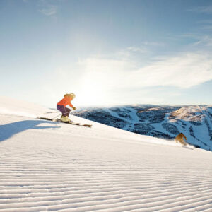 Snow sure slopes: where to head for late season skiing Skier in an orange jacket and purple trousers descends a groomed slope under bright sun, with snow-covered mountains stretching across the horizon.