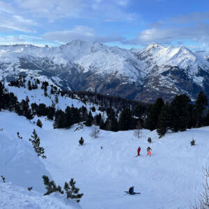 Two skiers descend a snow-covered slope dotted with pine trees, overlooking a vast range of snow-capped mountains under a partly cloudy blue sky.