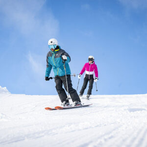 New La Mega Bell Easter Event comes to La Plagne, France Two skiers in helmets and goggles carve down a groomed slope under a bright blue sky.