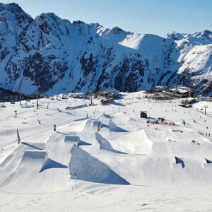 Spring skiing in the Austrian Tyrol A terrain park with sculpted snow jumps and ramps sits beneath towering snow-capped mountains, dotted with skiers and chairlifts under a clear blue sky.
