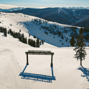 Make the most of spring skiing at Vail Resorts A snowy mountain vista with a wooden sign reading "THE LEGENDARY BACK BOWLS" framing a wide, treelined ski run.