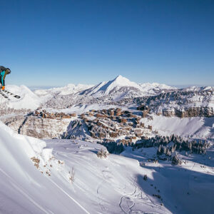 Early booking offers for 2025/26 on catered chalets in the French Alps Snowboarder launching off a powdery ridge above a snow-covered valley village, with sunlit mountain peaks stretching to the horizon under a clear blue sky.