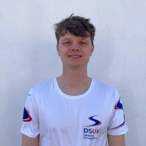 Duo To Run London Marathon for Disability Snowsport UK Smiling young man with dark wavy hair wearing a white DSUK Disability Snowsport UK t-shirt against a pale wall.