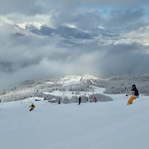 Skiers descend a snow-covered slope with cloud-shrouded mountains and frost-dusted trees in the background.