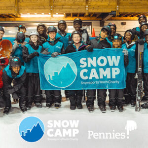 Charity challenge to ski all five UK indoor snow centres in a day A group of people in ski helmets and teal jackets pose together holding a 'Snow Camp Snowsports Youth Charity' banner indoors.
