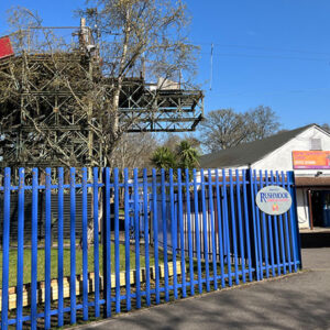 Alpine Snowsports Centre set to re-open A blue metal fence with a Plushmoor sign stands before a white building and a towering metal ski lift structure under a clear blue sky.