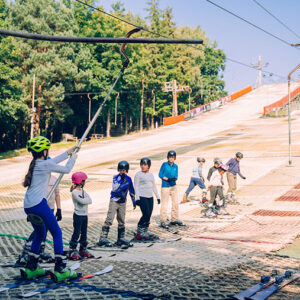Date set for Alpine Snowsports Centre re-opening Children in helmets on skis line up at a button lift on a dry ski slope matting, with an instructor guiding them beside sunlit trees.