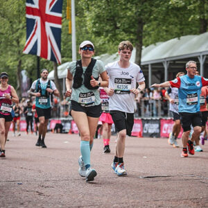 Runners wearing numbered race bibs jog past Union Jack flags along a tree-lined city street during a marathon event.