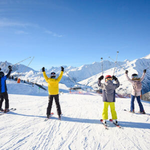 2025-26 UCPA family ski holidays now on sale Four skiers raise their poles triumphantly on a sunlit snow slope beneath snow-capped mountain peaks.