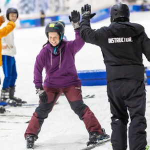 Snowsport England launches Way2Snow: affordable real snow ski lessons Smiling skier in a purple jacket high-fives an instructor on an indoor snow slope.