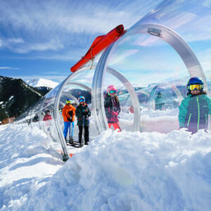 Children in ski gear gather beside a transparent acrylic tunnel on a snowy mountain slope under a bright blue sky.