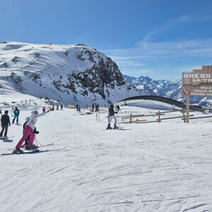 Hop aboard a magical chairlift in the Italian Alps Skiers gather on a snowy slope beneath a multilingual welcome sign for Gressoney La Trinité, with rugged, snow-capped mountains rising in the background.