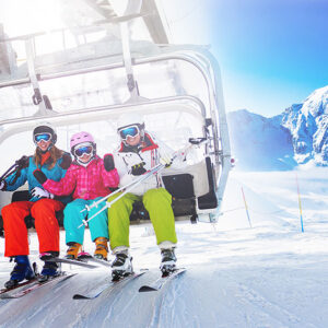 Three skiers in colourful jackets, helmets and goggles ride a chairlift with snow-capped mountains behind.