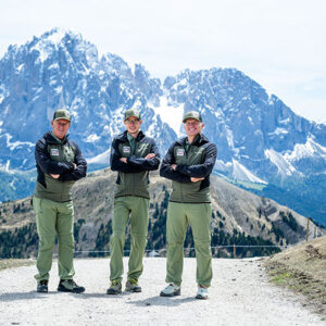 Three men in matching green uniforms and caps stand with arms folded on a mountain path before a towering, snow-dusted rocky peak.