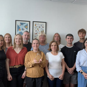 A group of around twelve people pose together in an office, with the central person holding a clear award, beneath two framed winter-themed artworks.