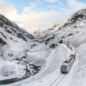 A train winds through a snow-covered mountain valley alongside a partially frozen river, with frost-laden trees and rocky peaks rising on either side.