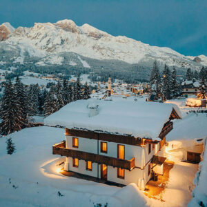 A snow-covered alpine chalet with warmly lit windows sits before towering snow-capped mountains at dusk.