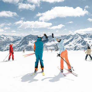 Two skiers high-five on a sunny mountain slope while others ski nearby, with snow-capped peaks and a cloudy blue sky in the background.