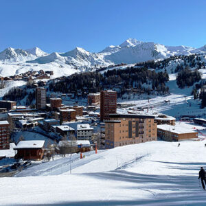 Snow-covered alpine resort with apartment blocks, distant peaks, and a lone skier descending a groomed slope.
