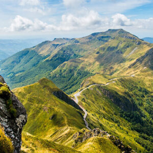 Lush green mountain ridges with a winding footpath stretch into the distance under a partly cloudy sky, with a rocky outcrop framing the foreground.