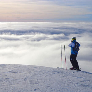 Why Solo Skiing is Having a Moment in 2025/2026 A skier in a blue jacket stands on a snowy ridge above a vast sea of clouds, gazing out across the horizon.