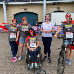 Travel agent partners with Paralympian to raise money for DSUK Five women wearing red superhero masks, capes, and "Superhero Series" t-shirts pose together, with one in a wheelchair and one beside a bicycle.