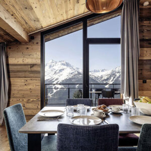 A wooden alpine dining room with a set table and breakfast spread, framed by large windows revealing snow-capped mountains.
