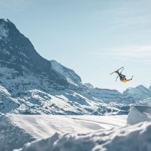 Jungfrau Ski Region joins the new AlpsPass A skier in a yellow jacket performs a mid-air trick above a snow jump, with snow-capped mountains behind.