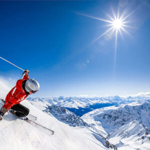 Jet2.com launches first ski programme from Luton Skier in a red jacket and white helmet carves down a sunlit snowy slope against a deep blue sky and distant mountain range.