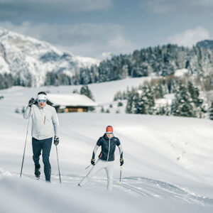 Wildtrack Nordic – Elevating the Art of Cross-Country Skiing Two cross-country skiers in white and grey winter kit ski across fresh snow towards the camera, with a wooden chalet and pine-covered slopes behind.