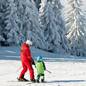 Ski tour operator reveals its top 10 family-friendly resorts An adult in a red ski suit and white helmet helps a small child in a green jacket and blue helmet learn to ski on a snowy slope backed by frost-covered trees.