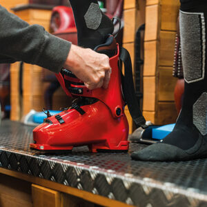 WIN a £500 voucher from The Skiers Lounge Hands fasten the buckles of a bright red ski boot worn with black and grey ski socks, standing on a metal platform.