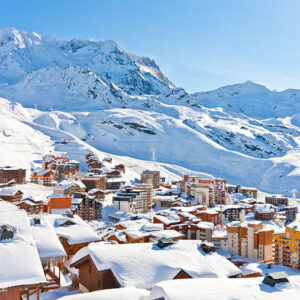 Chalet company shares top ski holiday choices for this winter Snow-covered alpine resort village with multi-storey buildings and chalets nestled beneath towering, sunlit mountain peaks under a clear blue sky.