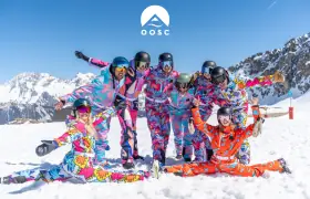 Group of skiers and snowboarders in vibrant matching onesies pose together on a snowy mountain beneath a clear blue sky.