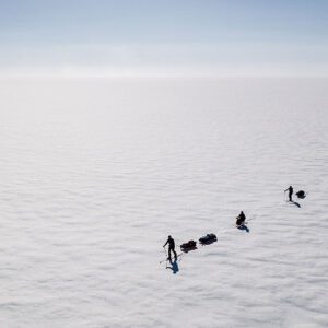 Three skiers in silhouette haul pulks in single file across a vast, flat snowfield under a pale hazy sky.