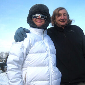 The real Minty Clinch – a skiing legend Two people pose together in the snow, one bundled in a white puffer jacket with a fur hat and ski goggles, the other in a dark jacket with an arm around them.