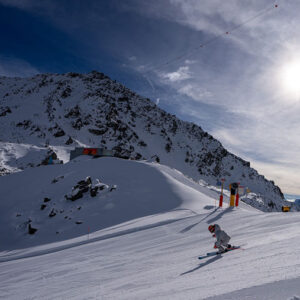 Verbier Kicks Off Ski Season Four Weeks Early A skier in a white jacket carves down a sunlit snow slope with a rocky mountain and ski lift cables in the background.
