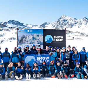 Snow Camp invites UK Businesses to Join AJ Bell 3 Valley Rally 2026 A large group of people in blue jackets and ski goggles pose on snow in front of a "Snow Camp" banner with mountains behind.