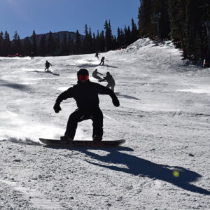 Colorado's 2025-26 ski season gets underway Snowboarder in dark clothing and goggles carving down a sunny, tree-lined piste with other skiers in the background.
